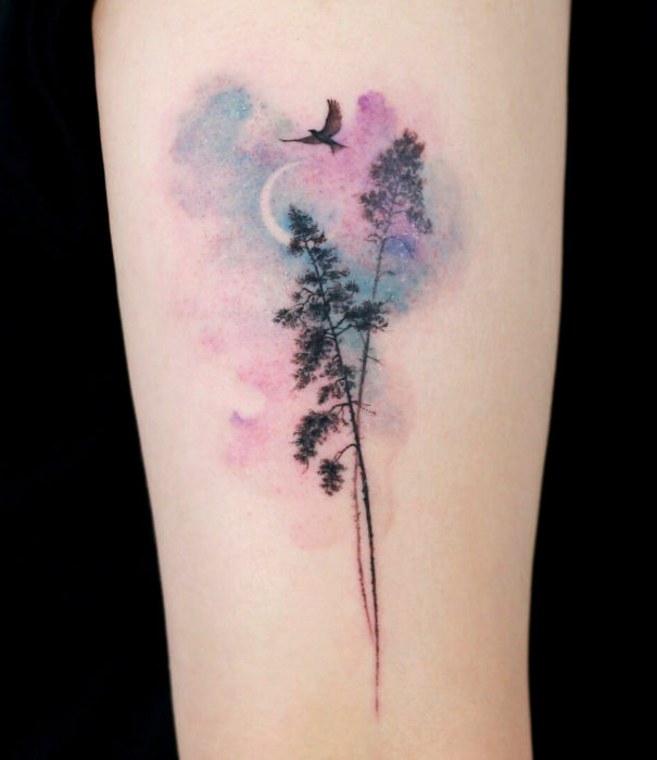 Diseños bonitos de tatuajes de acuarelas; tatuaje de árboles con universo de fondo en colores pastel en el brazo