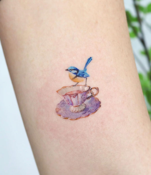 Diseños bonitos de tatuajes de acuarelas; tatuaje de pájaro azul en taza vintage de té