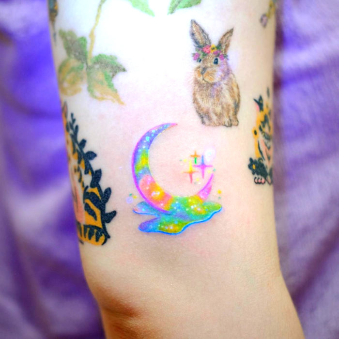 Tatuadora hace tatuajes bonitos, delicados y femeninos que brillan; tatuaje de media luna de colores morado, azul, rosa y verde pastel, conejo, en el brazo