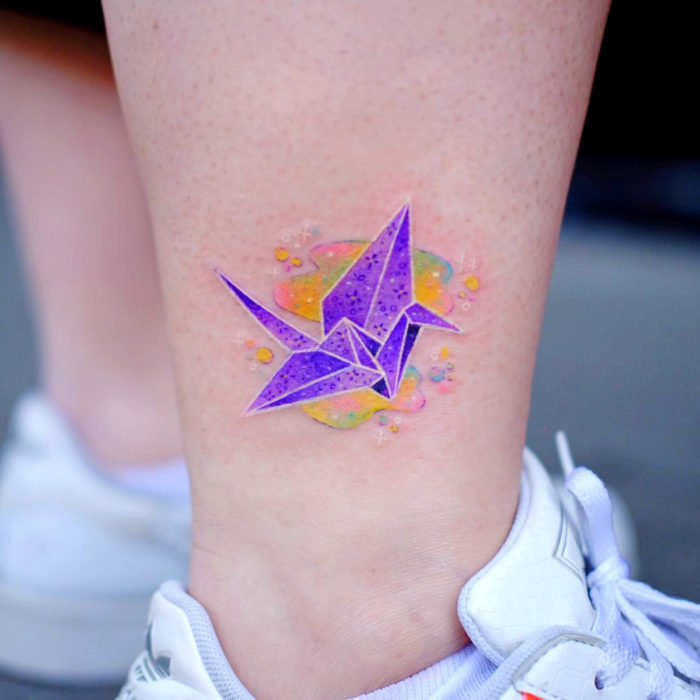 Tatuadora hace tatuajes bonitos, delicados y femeninos que brillan; tatuaje de grulla de origami color morado en el tobillo