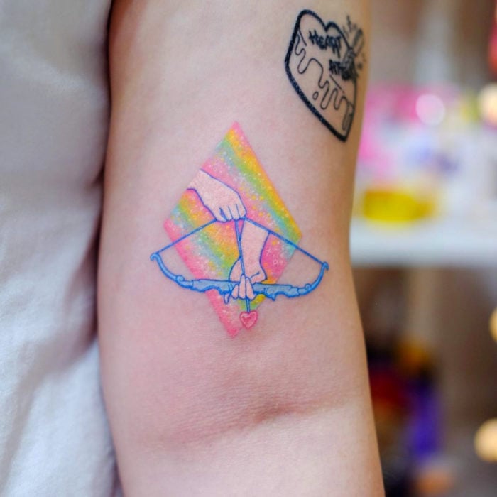 Tatuadora hace tatuajes bonitos, delicados y femeninos que brillan; tatuaje de arco con flecha de querubín, colores amarillo, rosa, verde y azul pastel en el brazo