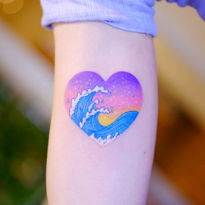 Tatuadora hace tatuajes bonitos, delicados y femeninos que brillan; tatuaje de ola japonesa dentro de corazón color morado, rosa, amarillo y azul pastel