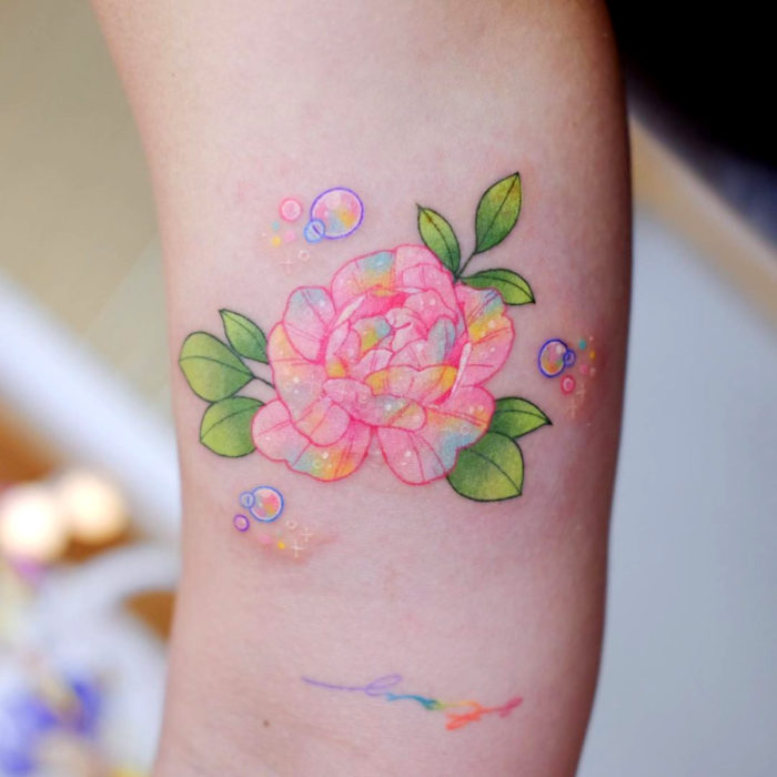 Tatuadora hace tatuajes bonitos, delicados y femeninos que brillan; tatuaje de flor peonia color rosa, amarillo y azul pastel en el brazo