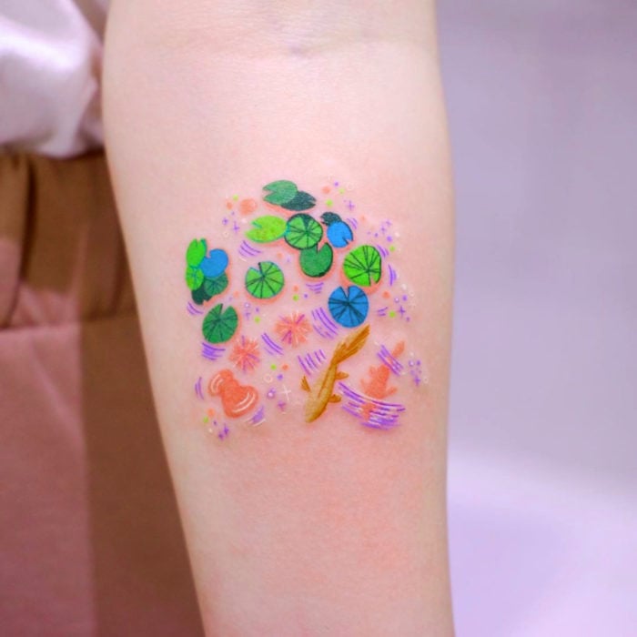 Tatuadora hace tatuajes bonitos, delicados y femeninos que brillan; tatuaje de hojas de nenúfar colores verde, azul y peces koi en el brazo