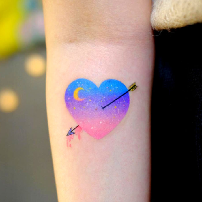 Tatuadora hace tatuajes bonitos, delicados y femeninos que brillan; tatuaje de corazón con flecha y Luna de colores azul, morado y rosa pastel en el brazo