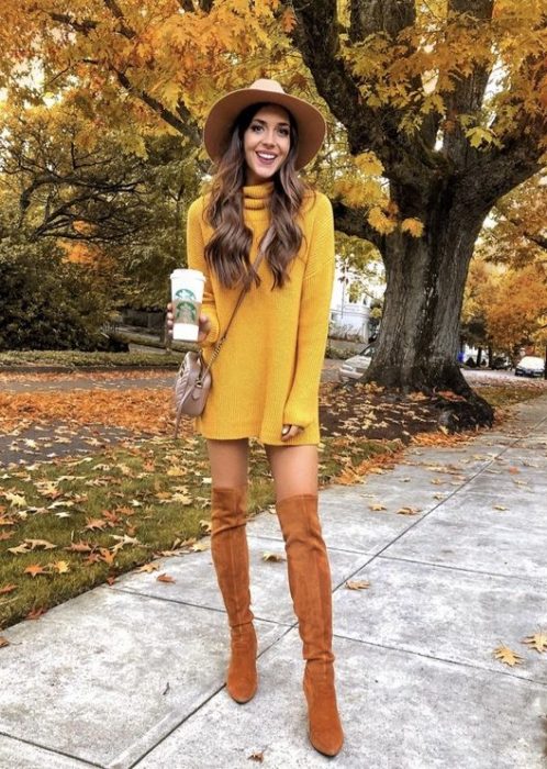 Chica delgada de cabello largo y ondulado con vestido amarillo de manga larga y cuello de tortuga con botas color camel y un sombrero