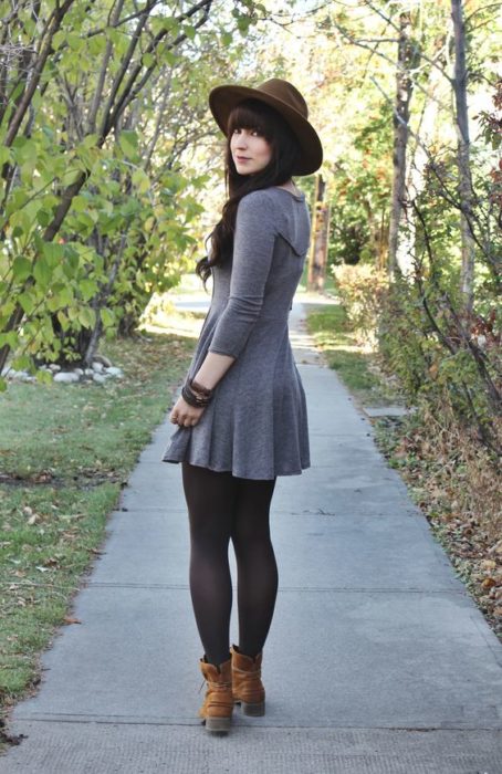 Chica delgada de cabello largo y oscuro con vestido gris y medias negras y sombrero