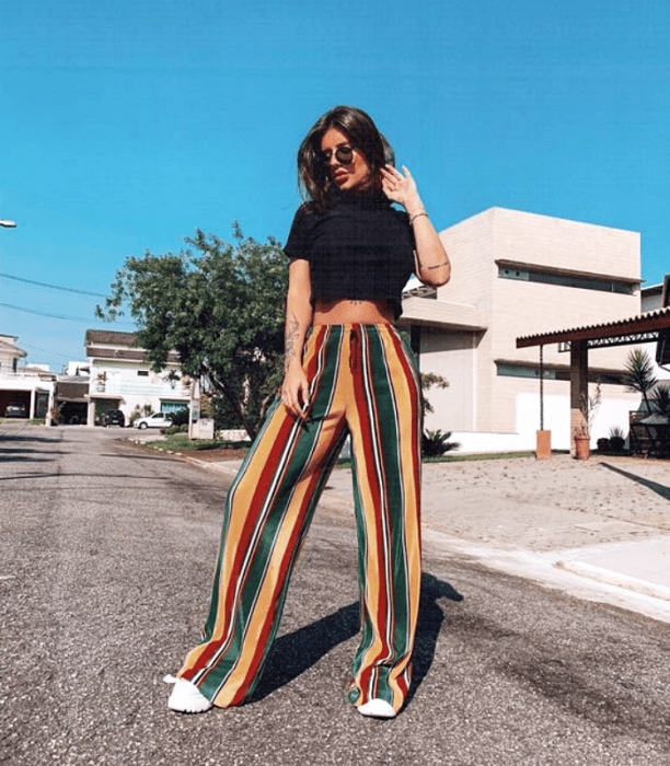 chica de cabello castaño corto con lentes de sol, camiseta negra y pantalones de rayas verdes, amarillas y rojas con tenis blancos