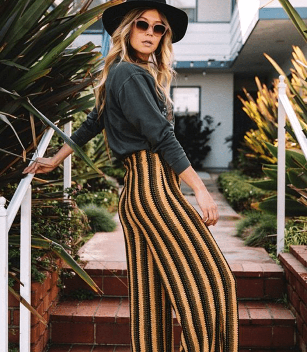 chica rubia con sombrero, lentes de sol, camiseta de manga larga gris con pantalones de rayas amarillas, verdes y negras