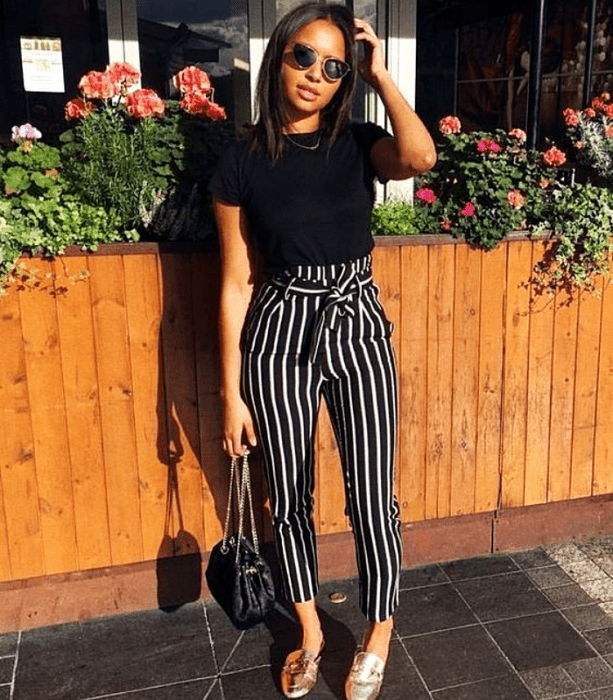 chica morena usando lentes de sol, camiseta negra y pantalones de rayas negras con blanco y flats doradas