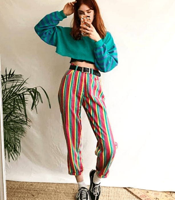 chica pelirroja usando un crop top de manga larga de color verde con lineas azules, pantalón de rayas amarillas, verdes, rojas, y blancas con tenis deportivos negros