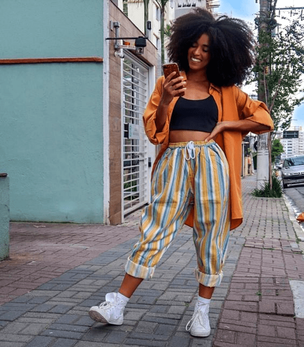 chica morena usando una camisa naranja con un crop top negro debajo y pantalones de rayas amarillo con azul y blanco y tenis deportivos blancos