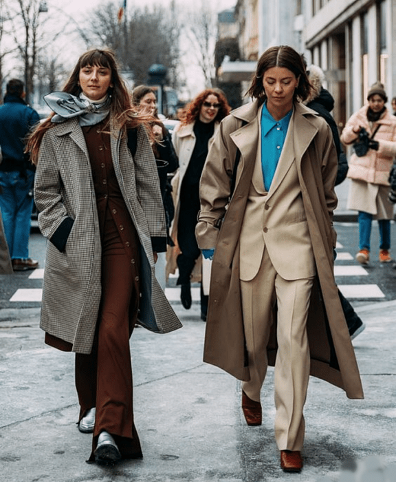 chica de cabello castaño usando una mascada azul, top de cuello alto blanco, saco café, pantalones de vestir cafés, abrigo blanco largo de cuadros y botines plateados, chica de cabello corto usando una camisa de vestir azul, saco beige, pantalones de vestir beige y abrigo largo café, botines cafés de punta cuadrada