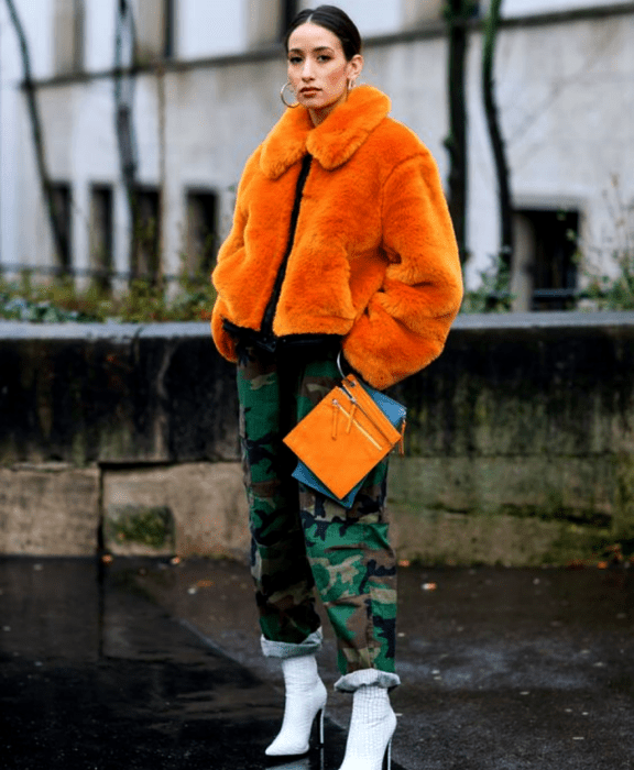 chica de cabello castaño usando un abrigo teddy naranja, chamarra negra debajo, pantalones de camuflaje, botines blancos de tacón y bolso naranja de mano