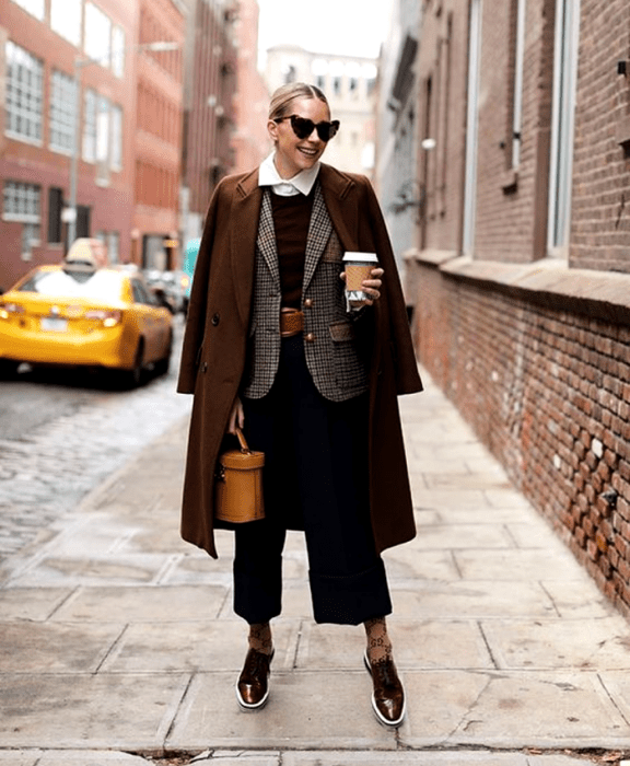 chica rubia usando lentes de sol en forma de corazon, camisa de vestir de cuello, suéter azul marino, cinturón café, saco café de cuadros, abrigo café largo, pantalones de vestir negros, calcetas cafés, zapatos cafés de vestir, bolso bucket naranja