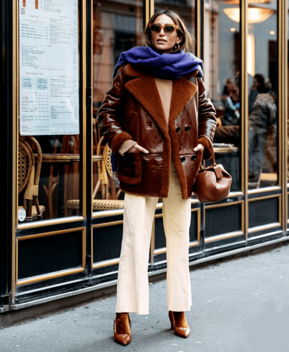 chica de cabello castaño usando lentes de sol, bufanda morada, abrigo café de piel, top beige, pantalones blancos, calcetas cafés y tacones cafés de piel, bolso café de mano