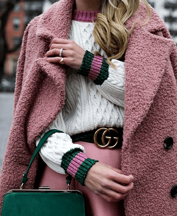 chica rubia usando un suéter blanco con cuello rosa y puños verdes con rosa, cinturón negro, falda rosa, abrigo teddy rosa y bolso verde de mano