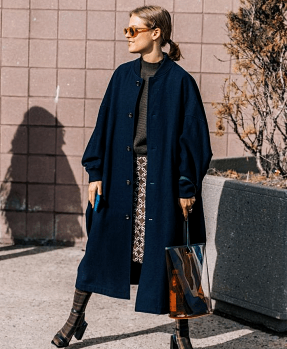 chica rubia usando lentes de sol, abrigo azul marino, suéter gris oscuro, falda con estampado de cuadros, bolsa transparente, calcetas cafés y tacones azul marino