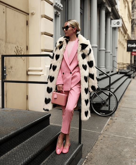 chica rubia usando lentes de sol, abrigo blanco con corazones negros, saco rosa, pantalones de vestir rosas, flats rosas y bolso rosa, top blanco debajo