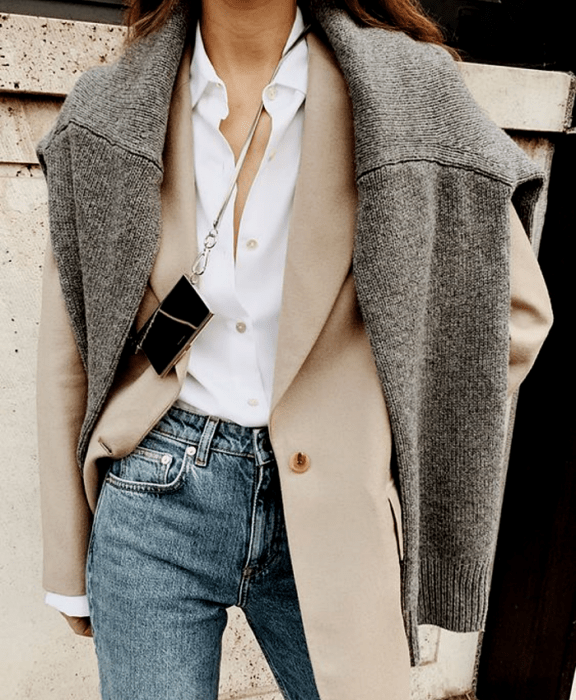 chica de cabello castaño usando un suéter gris, saco beige, camisa blanca y jeans a la cintura con bolso mini de metal 