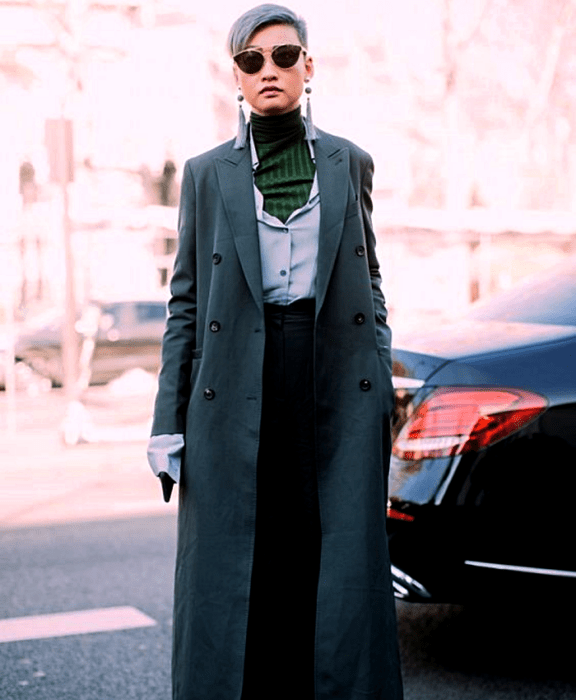 chica de cabello gris usando lentes de sol, top de cuello alto verde, camisa de vestir celeste, abrigo largo gris oscuro y pantalones de vestir negros