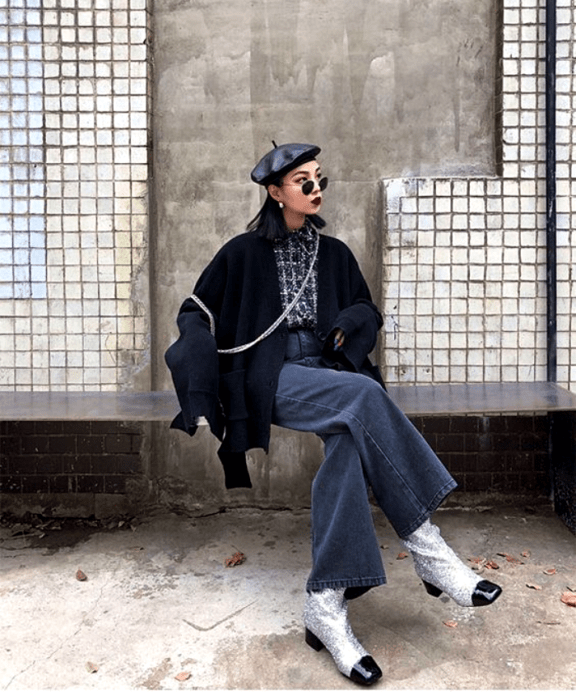 chica de cabello corto usando una boina, lentes de sol, camisa con estampado y cuello, cárdigan negro oversized, pantalones de mezclilla acampanados y botines plateados con diamantina y punta negra 