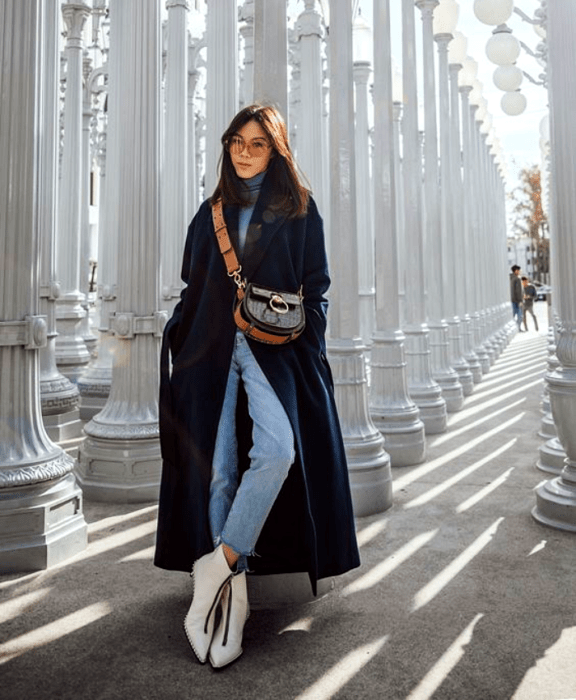 chica de cabello castaño largo usando lentes de sol, top de cuello alto color azul celeste, abrigo largo azul marino, jeans, botines blancos de cuero y bolso café