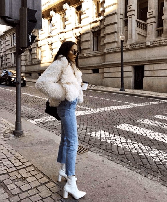 chica de cabello castaño largo usando lentes de sol, abrigo blanco tipo teddy, jeans ajustados, botines de cuero blancos con tacón y bolso negro