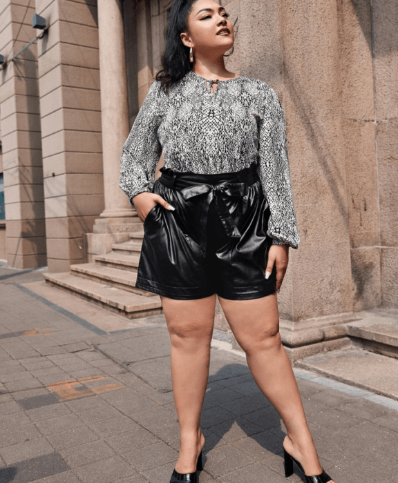 chica curvy morena usando blusa blanca con negro de manga larga, paperbag shorts negros de cuero y sandalias de tacón negras