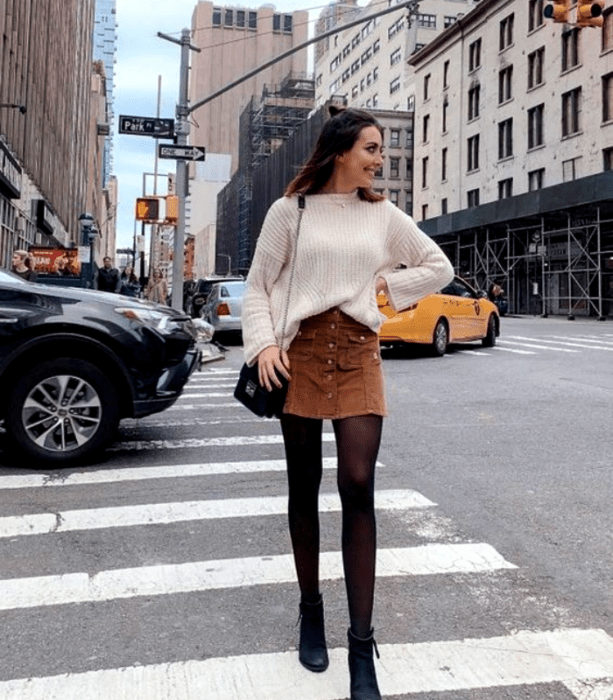 chica de cabello castaño largo con suéter blanco, falda café de pana, medias negras y botines negros y bolso pequeño