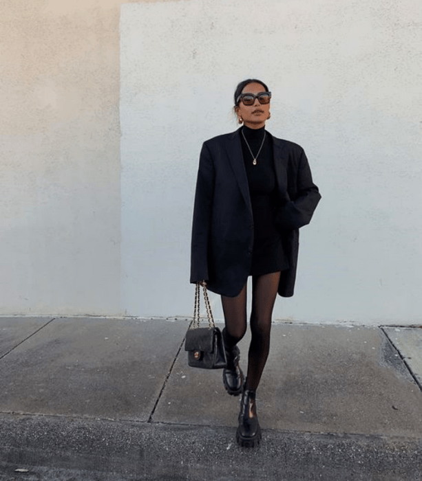 chica de cabello oscuro, lentes de sol, camiseta de manga larga, blazer oversized, medias negras, minifalda negra y botines negros 