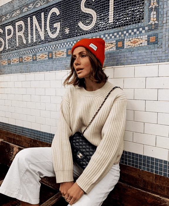 chica de cabello castaño usando un beanie naranja, suéter beige, bolso negro, pantalones blancos