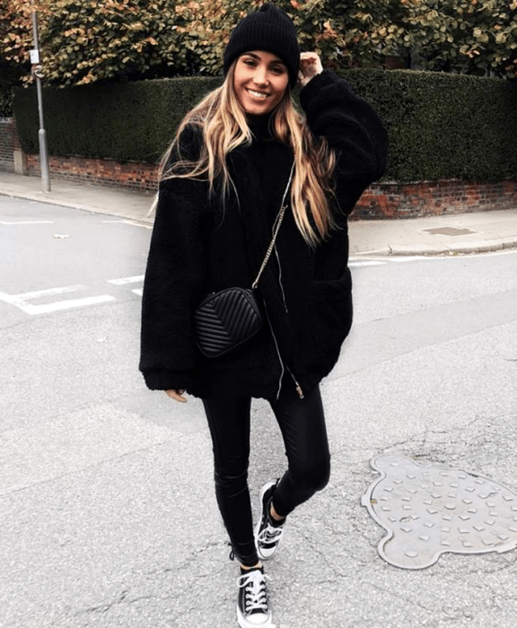 chica rubia usando un gorro beanie negro, abrigo oversized negro estilo teddy, leggings negros, tenis converse negros y bolso negro de piel