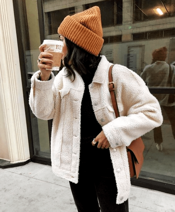 chica de cabello castaño usando un gorro beanie café, un abrigo teddy blanco, top negro de cuello alto y jeans negros, bolso café