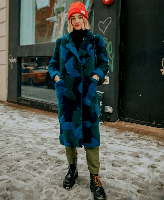 chica rubia usando un gorro beanie naranja, abrigo teddy azul con verde y negro, pantalones negros, botas negras de cuero de suela gruesa