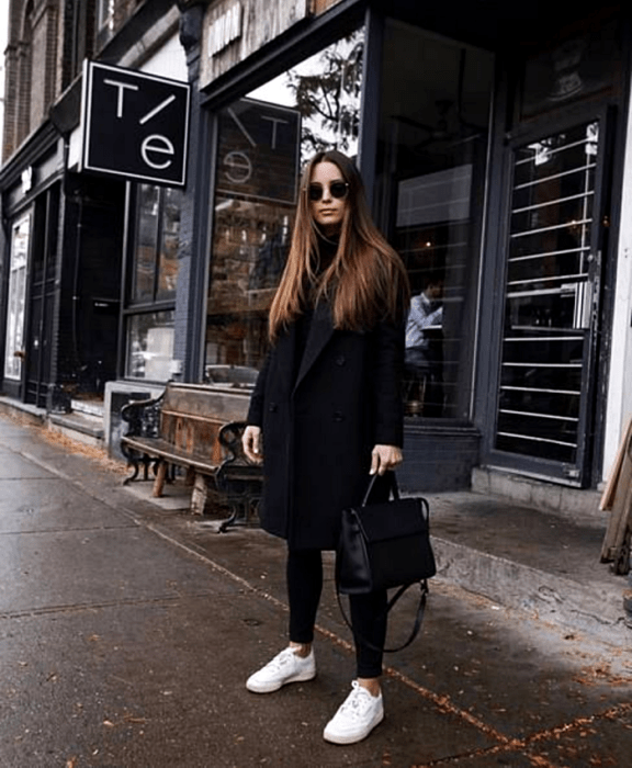 chica de cabello castaño usando lentes de sol, top negro de cuello alto, abrigo largo negro, leggings negros, tenis blancos converse y bolso negro