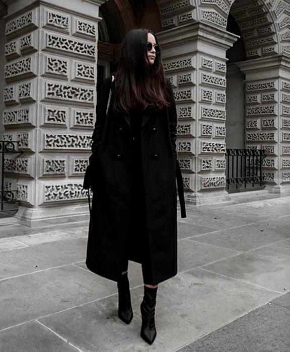 chica de cabello castaño usando lentes de sol, top negro, abrigo negro, skinny jeans negros, botines negros de tacón y bolso negro