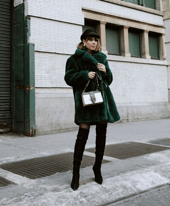 chica de cabello castaño usando un beret negro, abrigo largo verde de piel furry coat, botas largas negras de tacón, medias negras y bolso de mano blanco con negro