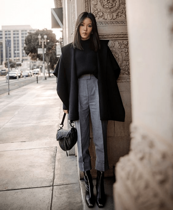 chica de cabello castaño largo usando un suéter negro, abrigo grande oversized negro, pantalones grises de vestir, botines negros de tacón y bolso negro de mano