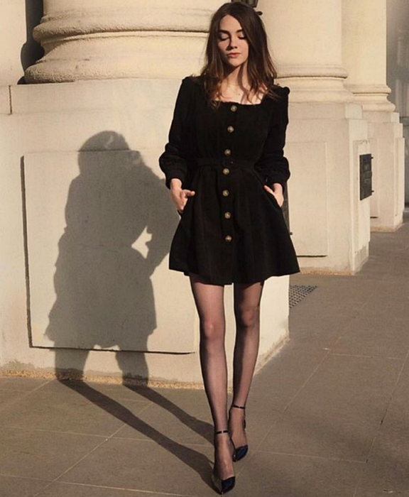 chica de cabello castaño usando un vestido de manga larga de pana negro con botones enfrente, medias negras y tacones negros con cintas en el tobillo