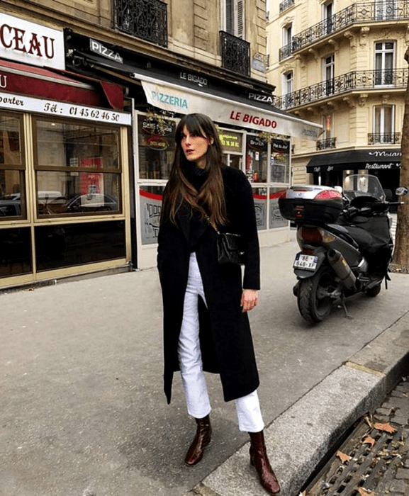 chica de cabello castaño usando un top negro, abrigo negro largo, pantalones rectos blancos, botines de cuero guindas y bolso negro pequeño