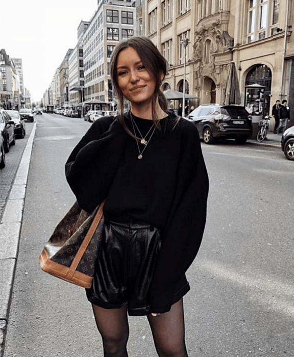 chica de cabello castaño usando un suéter negro, shorts cortos de cuero, bolso café y medias negras