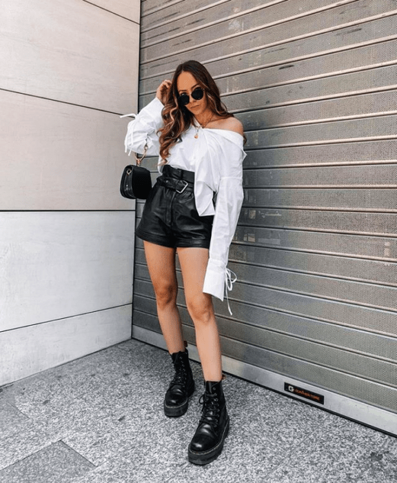 chica de cabello castaño largo usando lentes de sol, camisa blanca a los hombros, bolso pequeño negro, shorts cortos de cuero, botas de plataforma negras 