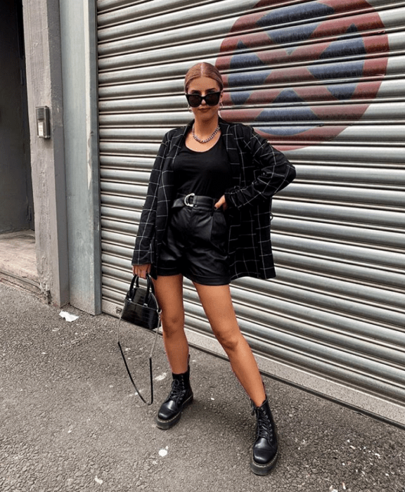 chica rubia usando lentes de sol, top negro, blazer saco con lineas blancas, shorts cortos de cuero, bolso pequeño de mano y botines negros de piso