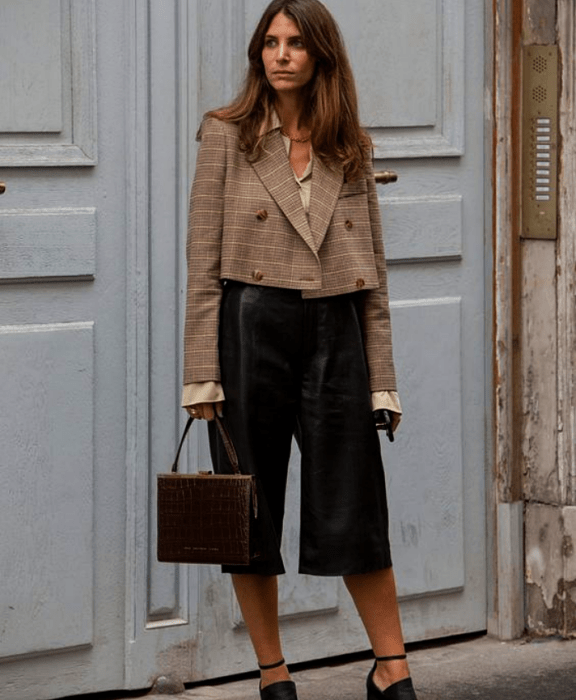 chica de cabello castaño claro largo usando una camisa beige, saco corto de cuadros, shorts tipo bermuda de cuero negro, bolso pequeño de cuero café y zapatos de tacón negros