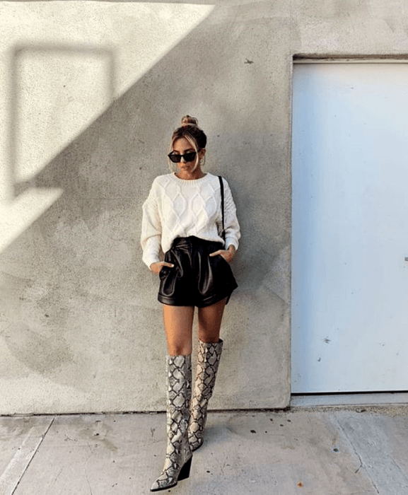 chica rubia con lentes de sol usando suéter blanco, shorts cortos de cuero, botas largas de estampado de serpiente y bolso negro pequeño