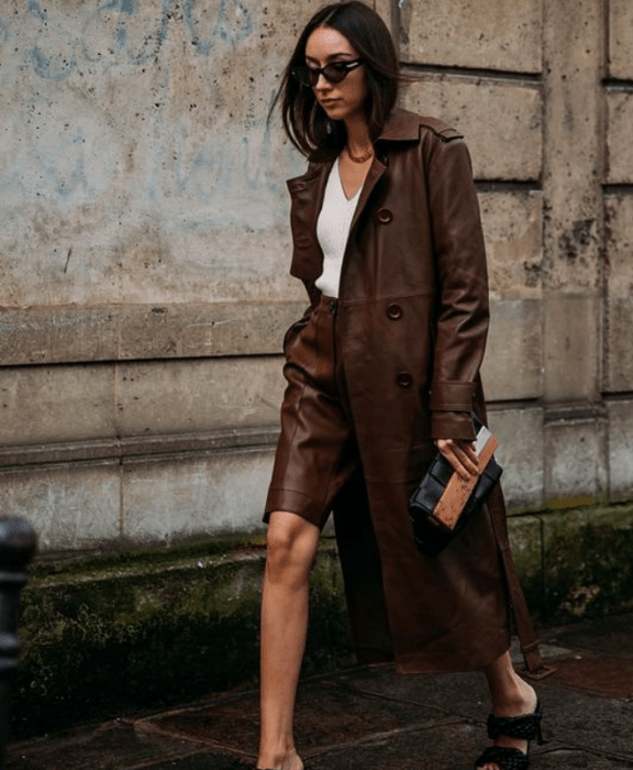 chica de cabello castaño usando lentes de sol, top blanco, gabardina de cuero café, shorts bermudas de cuero café y sandalias negras con tacón