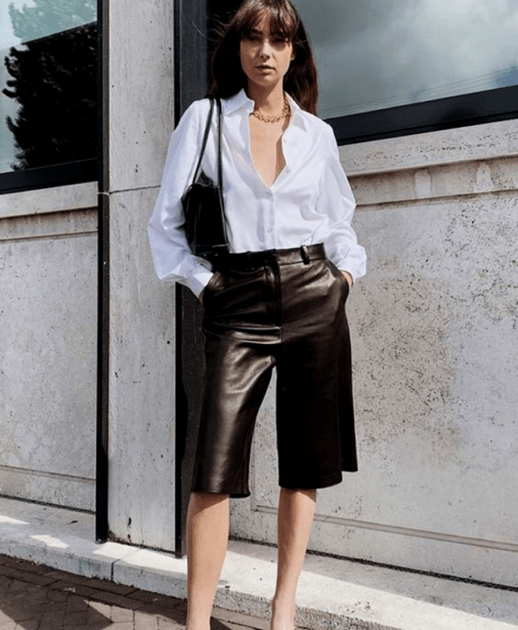 chica de cabello castaño largo usando una camisa de botones blanca con collar dorado, bolso pequeño negro de cuero, shorts tipo bermudas negras de cuero