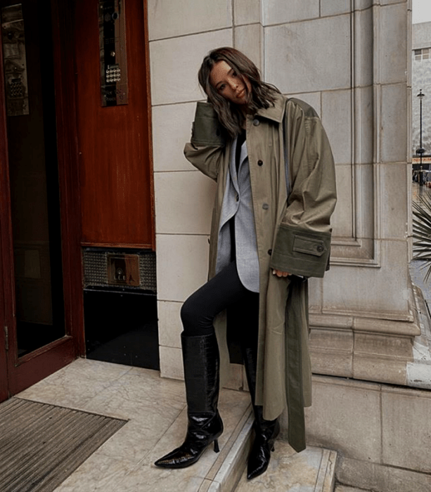 chica de cabello castaño largo usando un abrigo largo verde militar, un saco gris, leggings negros, botas largas de tacón negras