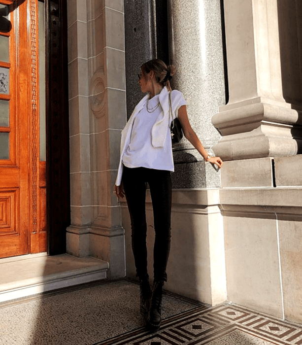 chica rubia con lentes de sol usando una camiseta blanca de manga larga, suéter blanco, leggings negros y botines negros de piso